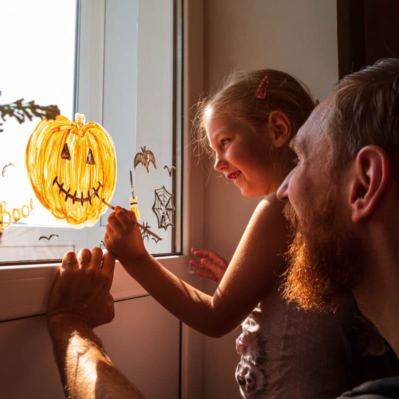 Girl painting pumpkin on window with her dad.